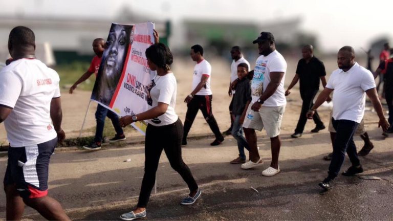 The procession moves through the streets around the National Stadium with LASTMA officers facilitating safe passage. Members and supporters carry the DNKI banner as traffic is managed around the group.